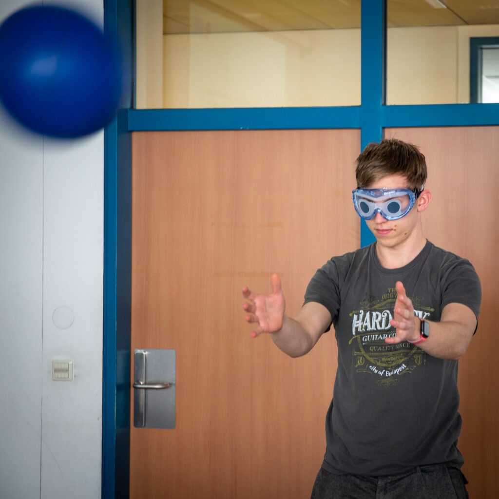 Ein Schüler mit aufgesetzter Simulationsbrille für Sehbeeinträchtigung versucht einen geworfenen Ball zu fangen