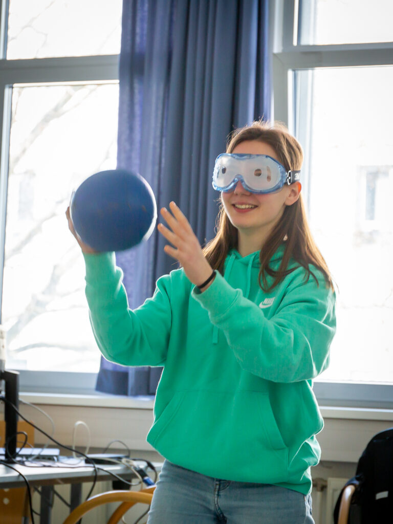 Eine Schülerin mit aufgesetzter Simulationsbrille für Sehbeeinträchtigung versucht einen Ball einem Mitschüler zuzuwerfen